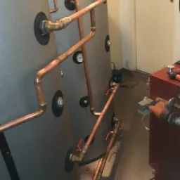 Dwa szare zbiorniki z miedzianymi rurami i zaworami, połączone w system. Widoczne manometry i elementy instalacji. Ujęcie z bliska, ukazujące detale połączeń.