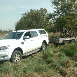 Geointec - Biały Ford Ranger 4x4 z wiertnicą na dwuosiowej przyczepie, zaparkowany w wysokiej trawie na słonecznym polu, z domem z czerwonym dachem w tle.