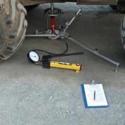 Geointec - Geotechniczne badanie nośności gruntu z użyciem hydraulicznego siłownika, ramy pomiarowej i pompy Parker pod kołem ciężkiej maszyny. Widoczny clipboard.