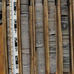 Rdzenie wiertnicze w drewnianej skrzynce, obok miarka. Widoczne warstwy skał i ich struktura. Próbki geologiczne z odwiertu.