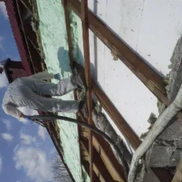 Pracownik w kombinezonie aplikuje piankę izolacyjną PUR na poddaszu, widoczne drewniane krokwie i fragment dachu. Błękitne niebo w tle, ujęcie z dołu.