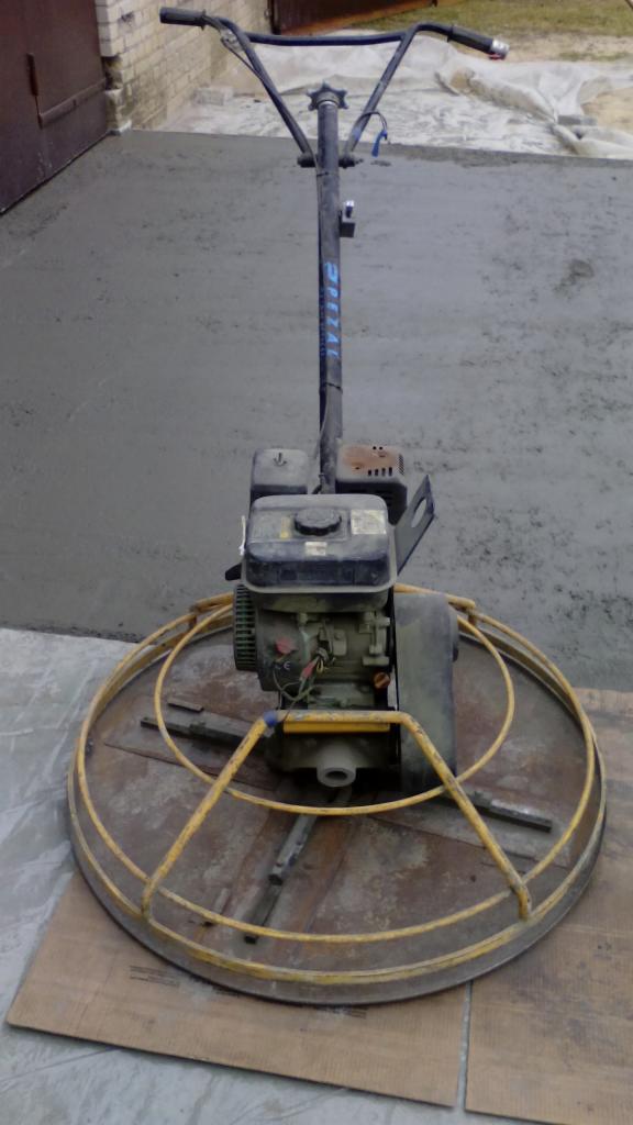 Zacieraczka mechaniczna na świeżej posadzce cementowej, z widocznymi śladami pracy i fragmentem ściany z cegły w tle. Sprzęt gotowy do użycia.