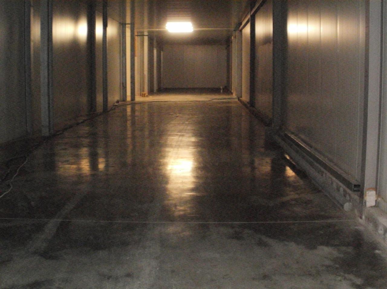 Długi, wąski korytarz z błyszczącą, gładką wylewką podłogową, odbijającą światło górnego oświetlenia. Ściany wykończone panelami, widoczne elementy konstrukcyjne.