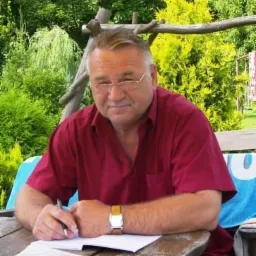 Mężczyzna w okularach, siedząc przy drewnianym stole w ogrodzie, pisze na kartce. W tle bujna roślinność.