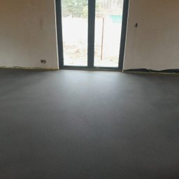 PHU JANUSZ MEDYŃSKI - Gładka, ciemnoszara posadzka w surowym wnętrzu z dużym oknem balkonowym. Widoczne zabezpieczenia ścian taśmą. Minimalistyczny design, naturalne światło.