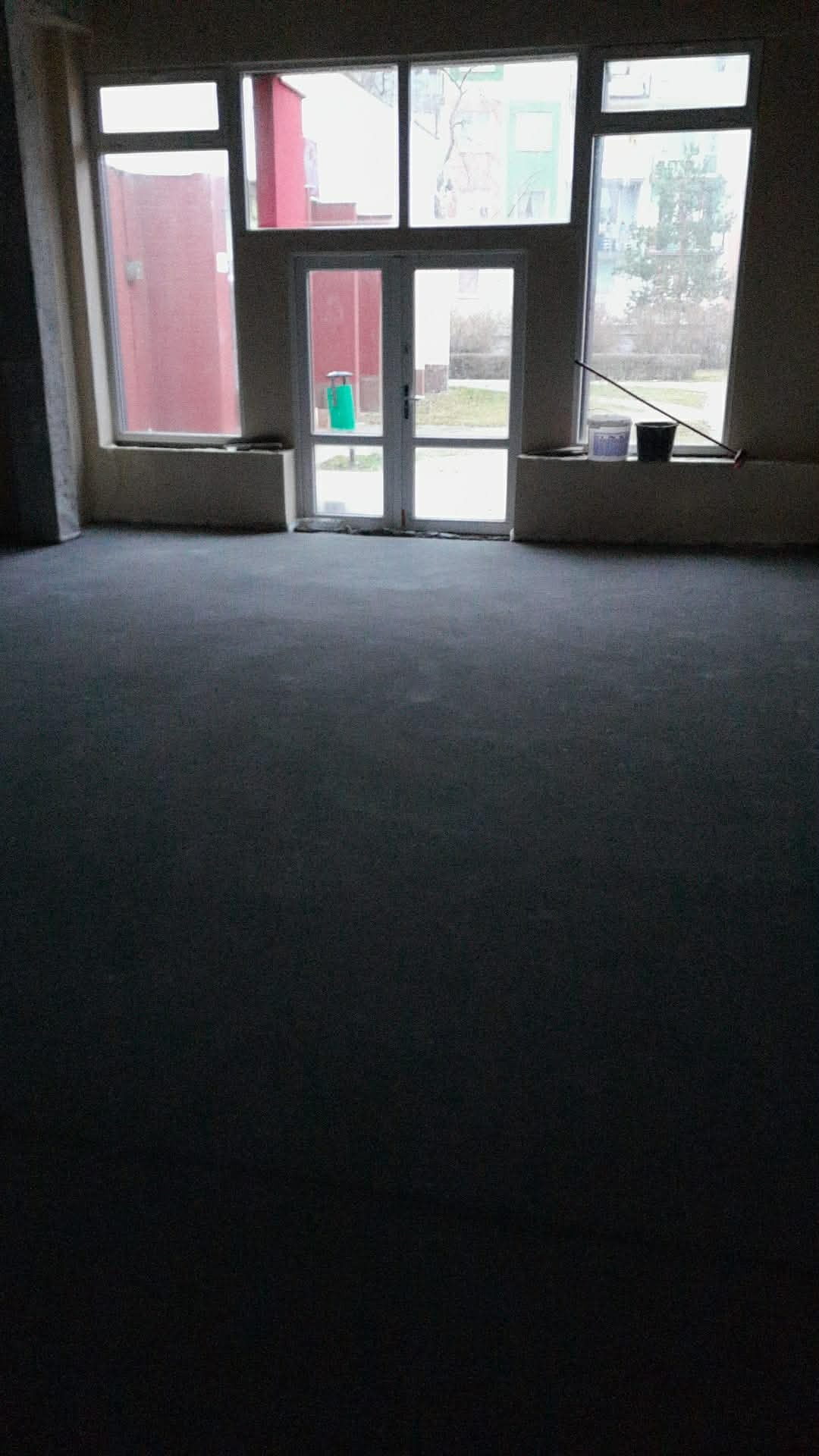 Wnętrze z szarą, świeżo wylaną posadzką jastrychową, widoczne okna i drzwi balkonowe. Na parapecie wiadro i miotła. Minimalistyczne wykończenie.