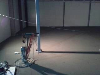 Wyrównana, szara posadzka w surowym wnętrzu, widoczna maszyna do zacierania z przewodami, oświetlona punktowo, zarys białej ściany w tle.