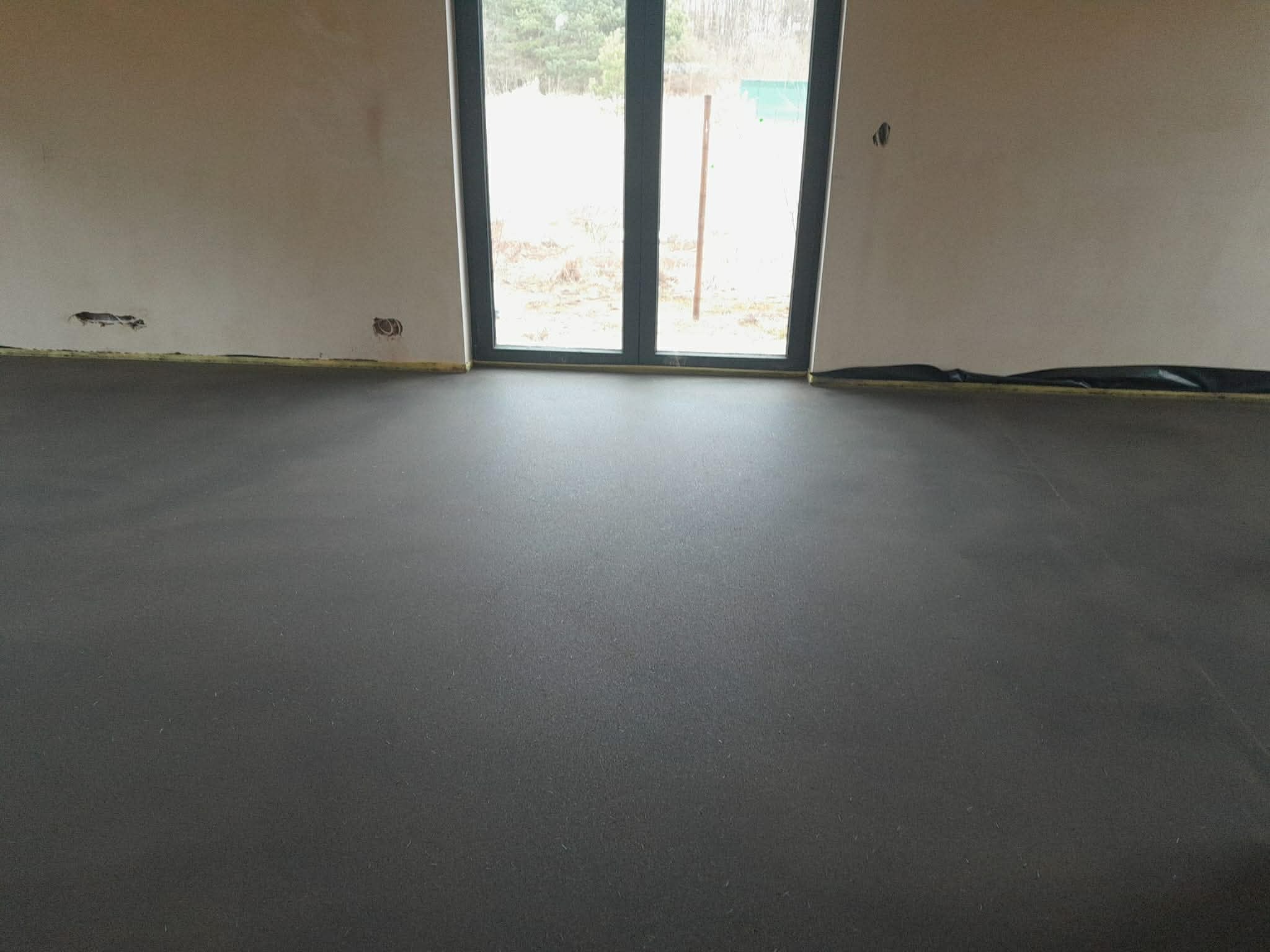 Gładka, ciemnoszara posadzka w surowym wnętrzu z dużym oknem balkonowym. Widoczne zabezpieczenia ścian taśmą. Minimalistyczny design, naturalne światło.