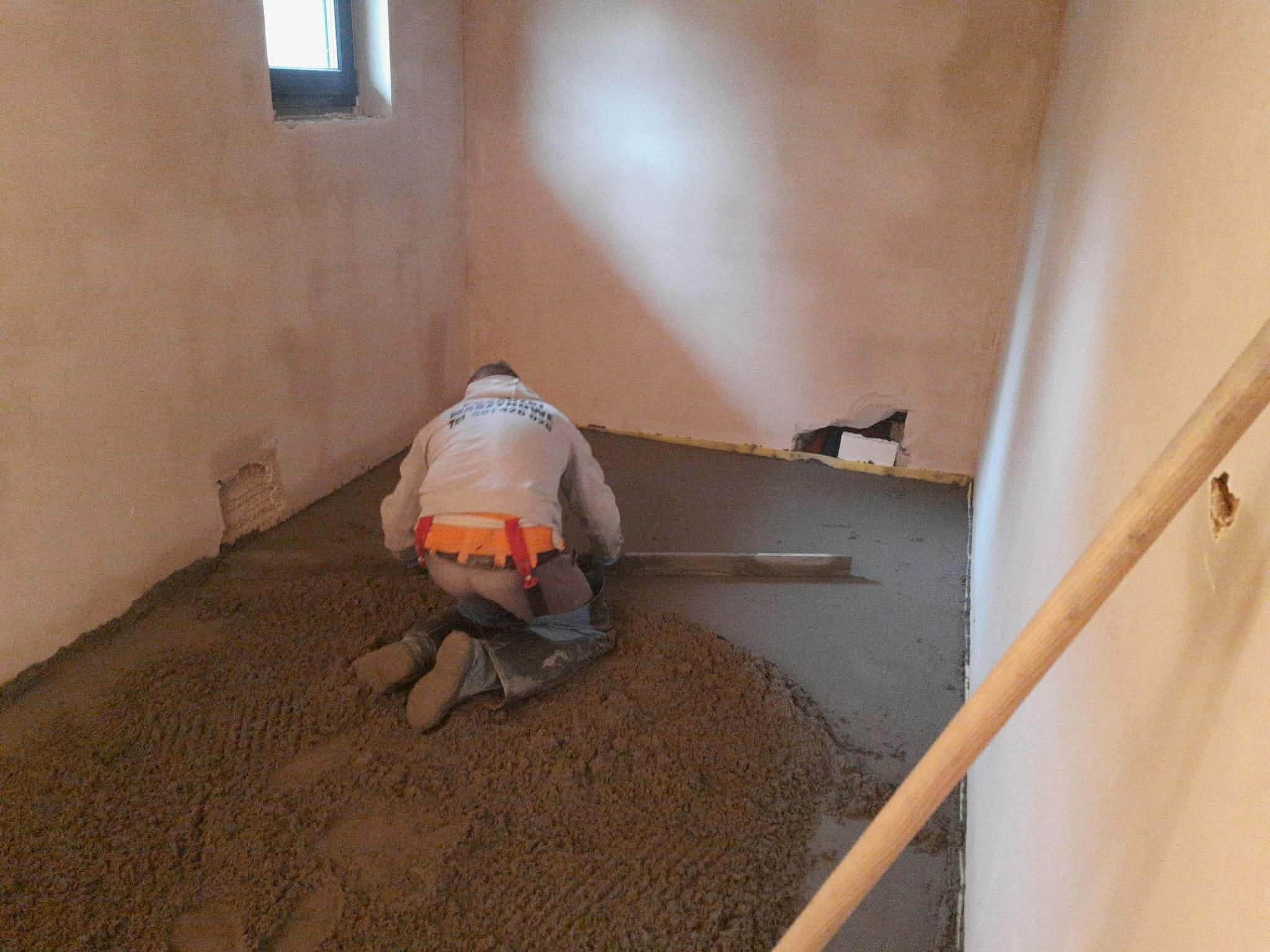 Pracownik w kombinezonie wyrównuje mokrą wylewkę jastrychową w pomieszczeniu z oknem. Widoczna łata i nierówności podłoża.