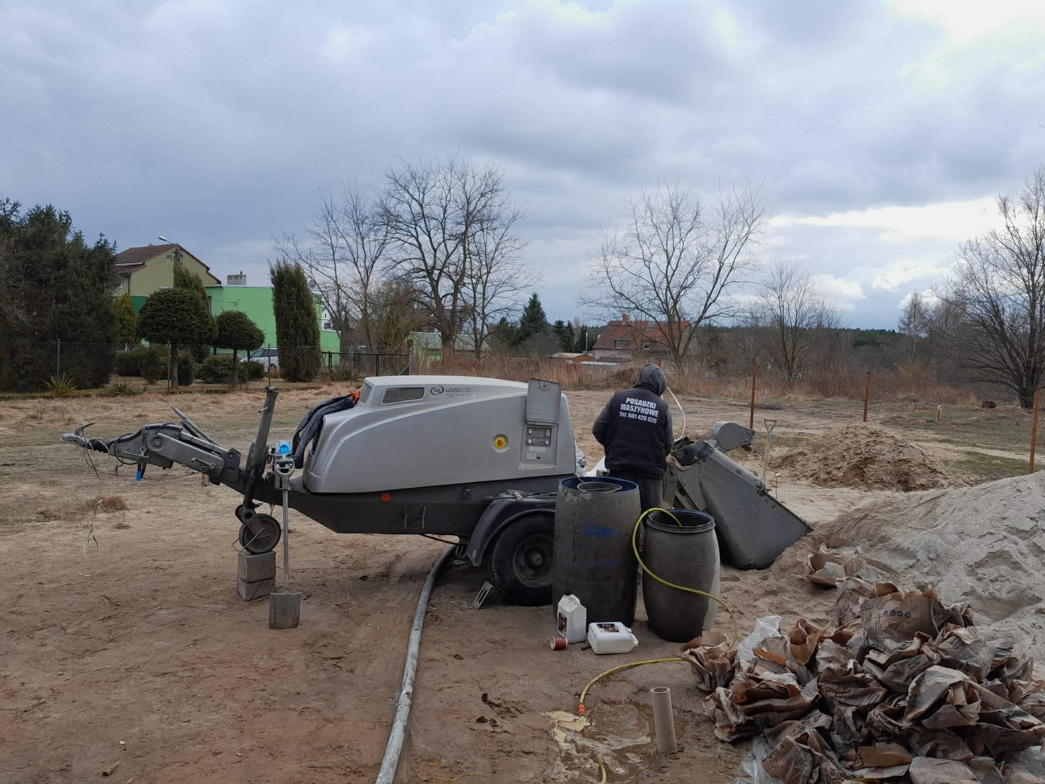 Maszyna do wylewania posadzek jastrychowych na budowie, widoczny pracownik, hałda piasku i puste worki po cemencie. Pochmurne niebo w tle.