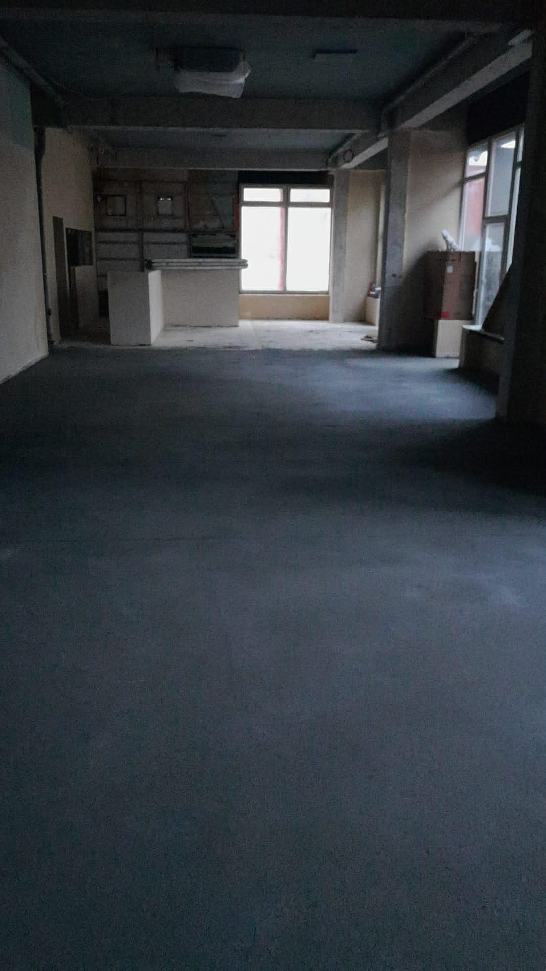 Wnętrze po wylaniu posadzki w szarym odcieniu. Widoczne okno z jasnym światłem dziennym i surowe ściany. Minimalistyczny, industrialny charakter przestrzeni.