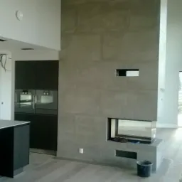 Nowoczesny kominek obłożony szarymi płytami w przestronnym salonie z drewnianą podłogą i zabudową kuchenną w tle. Widoczne narzędzia i wiadro.