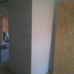 Wnętrze w trakcie prac wykończeniowych: narożnik z płyt gipsowo-kartonowych, widoczne ślady po wkrętach, płyta OSB na ścianie, przygotowanie do dalszych etapów prac.