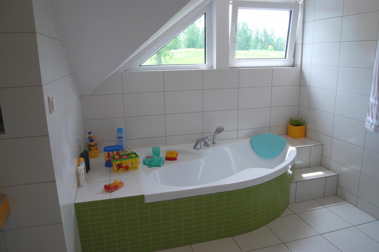 Nowoczesna łazienka na poddaszu z wanną obłożoną zieloną mozaiką, jasnymi płytkami i oknem dachowym z widokiem na zieleń. Dziecięce zabawki na wannie.