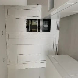 Nowoczesna, minimalistyczna kuchnia z białymi frontami szafek, srebrnymi listwami i wbudowanymi urządzeniami: piekarnikiem i mikrofalówką. Widoczne gniazdka elektryczne na ścianie.