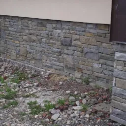 Kamienna elewacja budynku z kolumną, wykonana z nieregularnych szarych i beżowych kamieni, widoczna z bliska, z gruzem i roślinnością u podstawy.