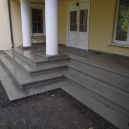 Wejście do domu z szarymi schodami kamiennymi, wsparte na kolumnach. Widoczne drzwi wejściowe i okno z kratkami. Żółta skrzynka gazowa po lewej stronie.