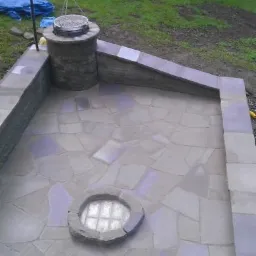 Wykończony taras z kamienną mozaiką i okrągłym świetlikiem; w tle trawnik i grill. Widoczne elementy kamienne i kompozycja z kamienia naturalnego.