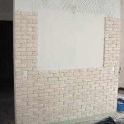 Złota Rączka - Sprawnie - Ściana z częściowo ułożoną dekoracyjną cegłą w jasnym odcieniu, widoczne gniazdko elektryczne i siatka pod tynk. W tle okno i pomieszczenie w trakcie remontu.