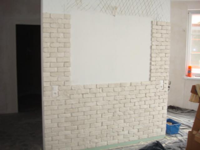 Ściana z częściowo ułożoną dekoracyjną cegłą w jasnym odcieniu, widoczne gniazdko elektryczne i siatka pod tynk. W tle okno i pomieszczenie w trakcie remontu.