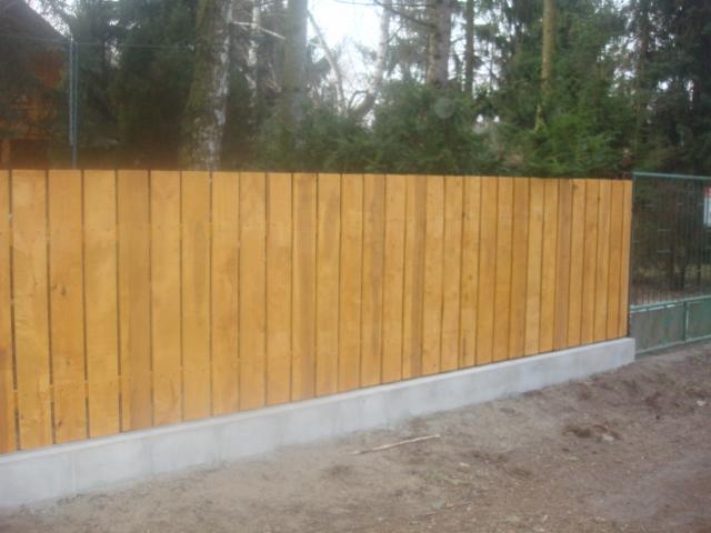 Nowoczesne ogrodzenie drewniane z podmurówką betonową, widok z zewnątrz, pionowe deski w naturalnym odcieniu drewna, brama w tle, drzewa.