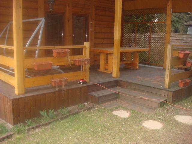 Drewniana altana z tarasem i schodami, ozdobiona wiszącymi donicami. Widoczne elementy ogrodu i trawnik. Jasne drewno konstrukcji kontrastuje z ciemniejszym tarasem.