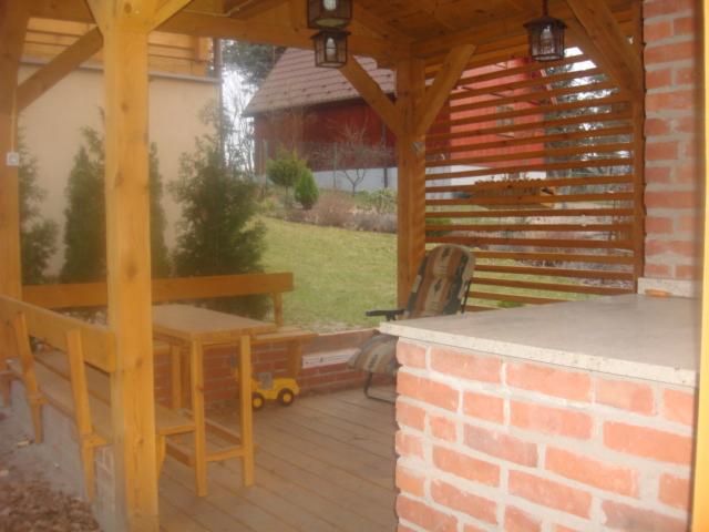 Drewniana altana z ceglanym grillem i stołem, widok na zielony ogród. Elementy drewniane w naturalnym kolorze, ceglany grill z blatem. Na podłodze deski.