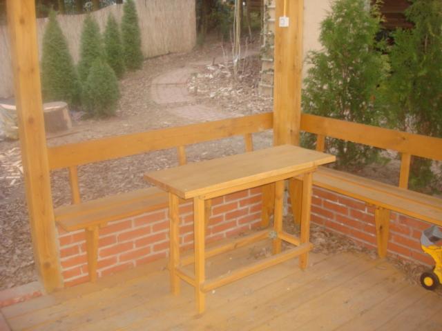 Drewniana altana z ławkami na ceglanej podmurówce i stołem. W tle widoczne tuje i ścieżka.