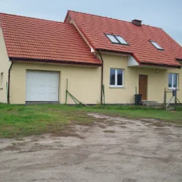 Żółty dom jednorodzinny z garażem i czerwoną dachówką w pochmurny dzień, widoczny fragment ogrodzenia i podjazd z piaskiem.