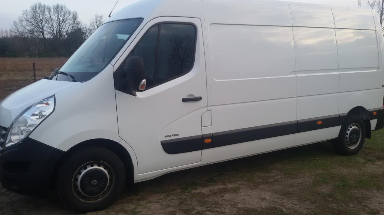 Biały bus Renault Master DCi 150 na tle pola i drzew, widok z boku. Pojazd zaparkowany na ziemi, widoczne opony i detale karoserii. Lekkie zachmurzenie na niebie.
