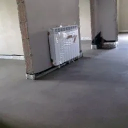 Gładka, szara wylewka w surowym wnętrzu z grzejnikiem owiniętym folią ochronną. Widoczne fragmenty ścian i kolumn w stanie deweloperskim.