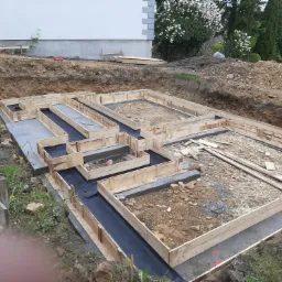 Drewniane szalunki przygotowane do wylania fundamentów domu, z widoczną hydroizolacją i wykopem ziemnym w tle.