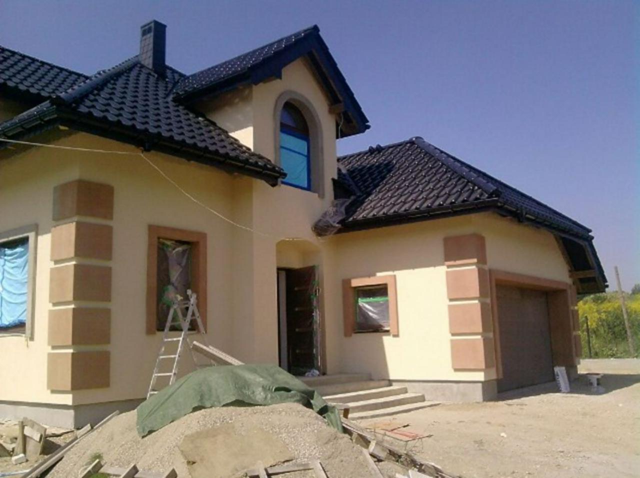 Niedokończony dom z beżową elewacją i ciemną dachówką. Okna zabezpieczone folią, widoczna drabina i materiały budowlane. Dom z garażem w trakcie budowy.