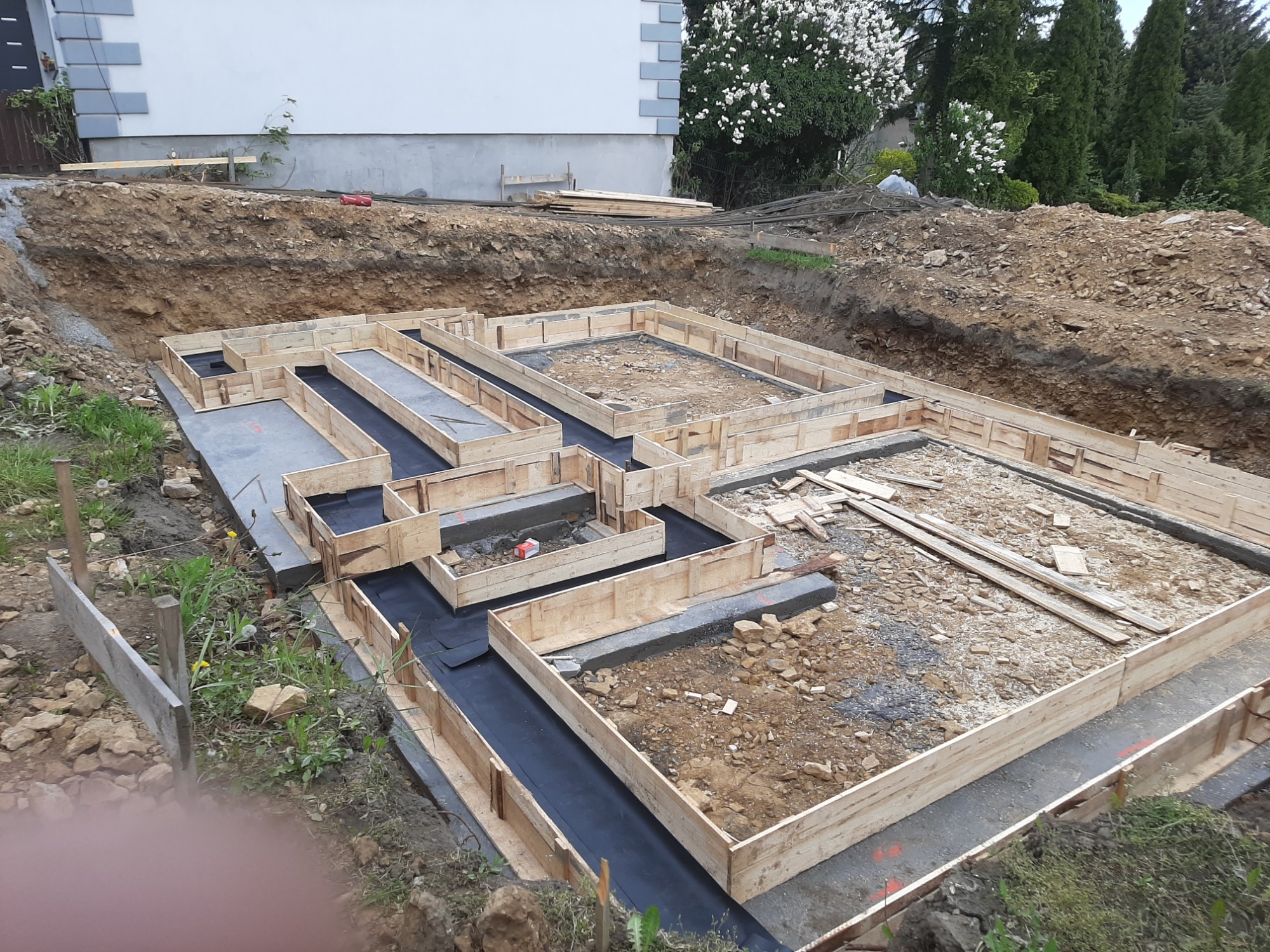 Drewniane szalunki przygotowane do wylania fundamentów domu, z widoczną hydroizolacją i wykopem ziemnym w tle.