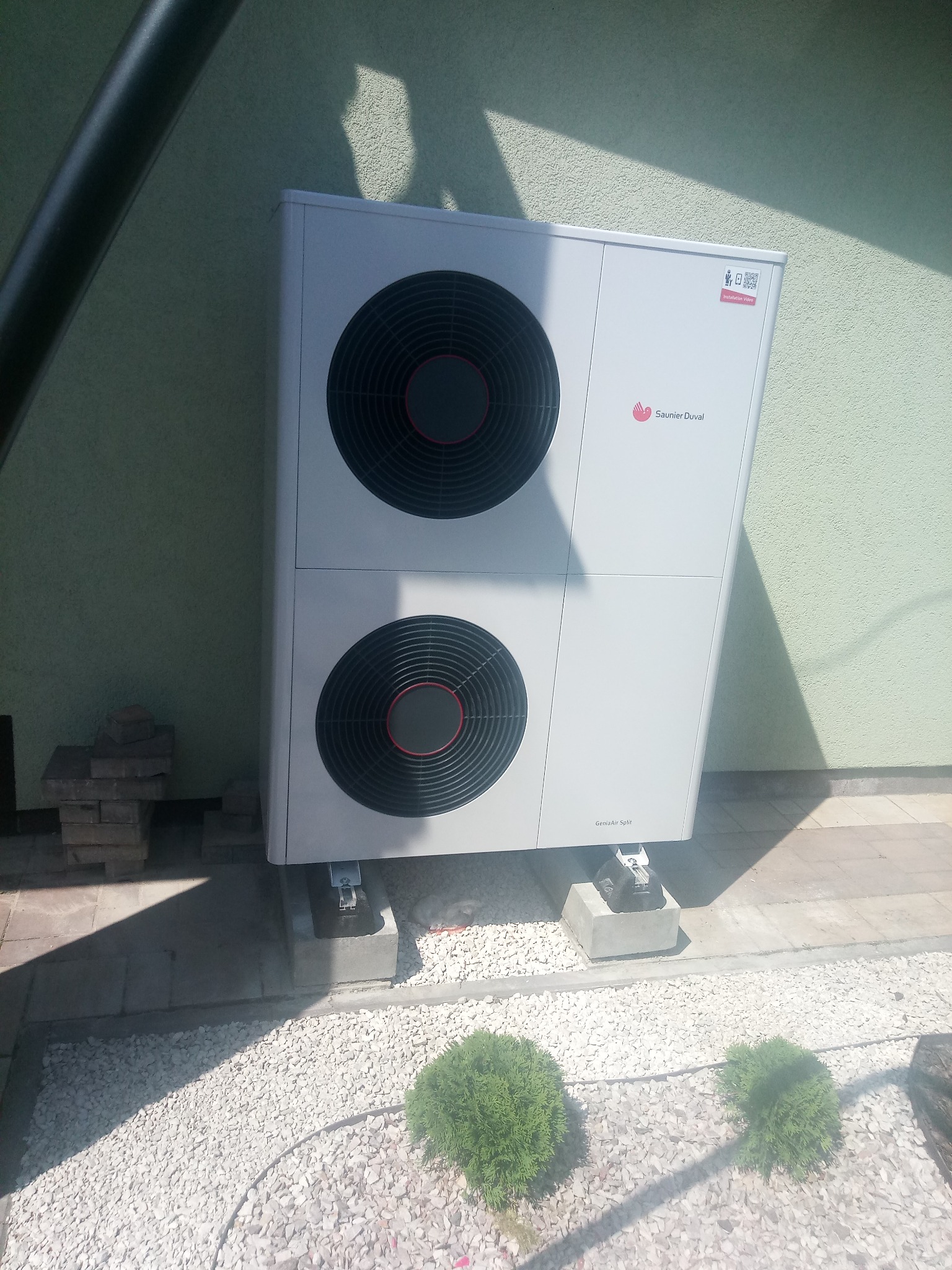 Biała pompa ciepła Saunier Duval GeniaAir Split zamontowana na betonowych podstawach, otoczona białym żwirem i zielonymi krzewami na tle zielonej ściany budynku.