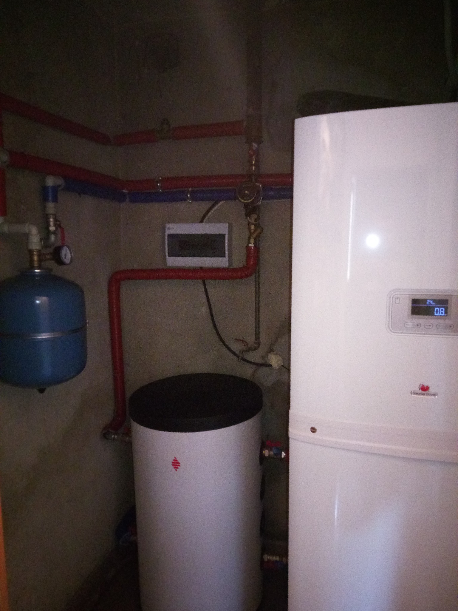 Biały piec grzewczy z wyświetlaczem cyfrowym, zbiornik akumulacyjny z czerwonym logo, niebieski zbiornik wyrównawczy i czerwono-niebieskie rury instalacji grzewczej na tle szarej ściany.