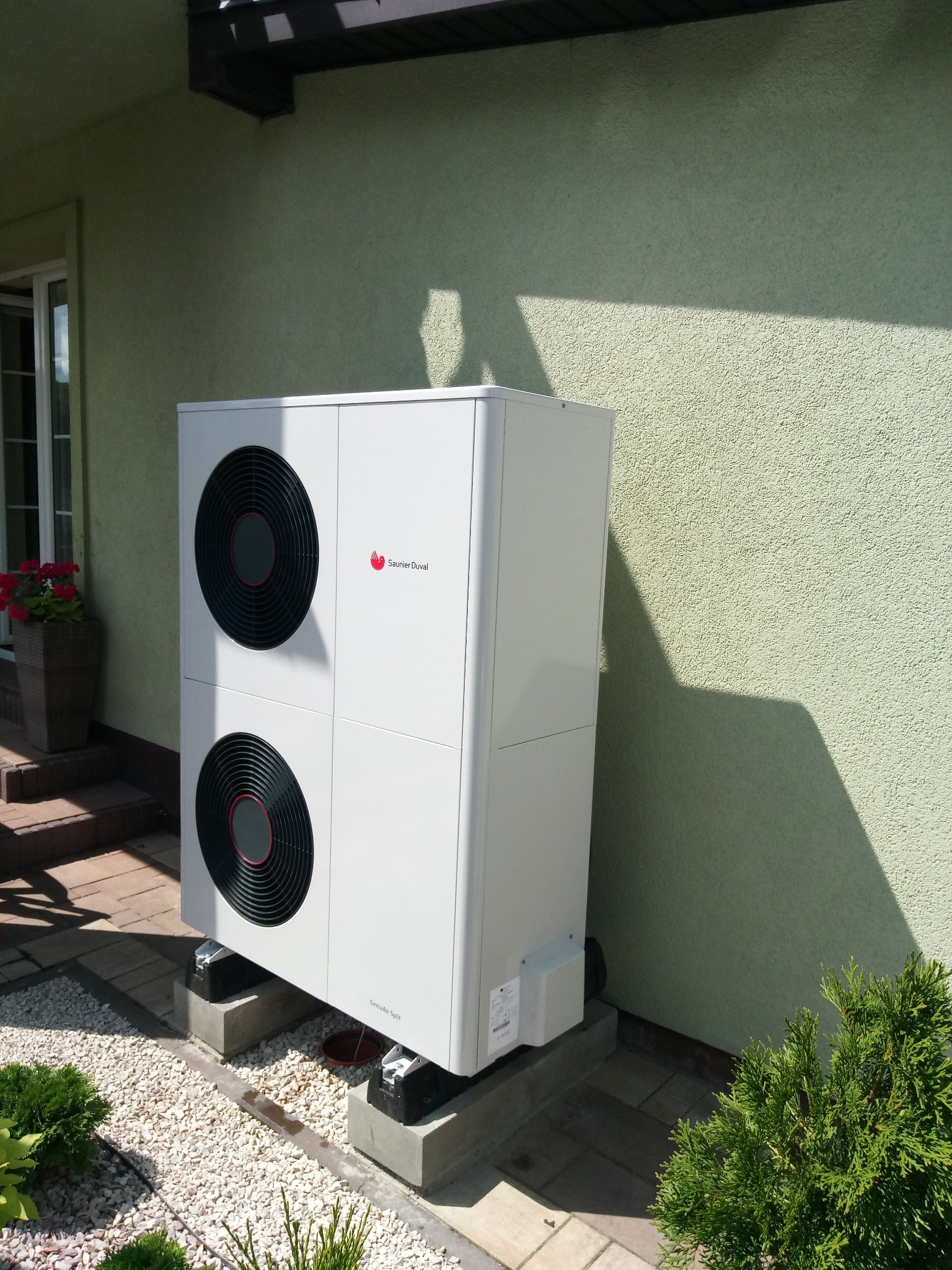 Biała pompa ciepła Saunier Duval GeniaAir Split zamontowana na betonowych podstawach przy zielonej ścianie domu, z widocznymi elementami instalacji i roślinnością w tle.