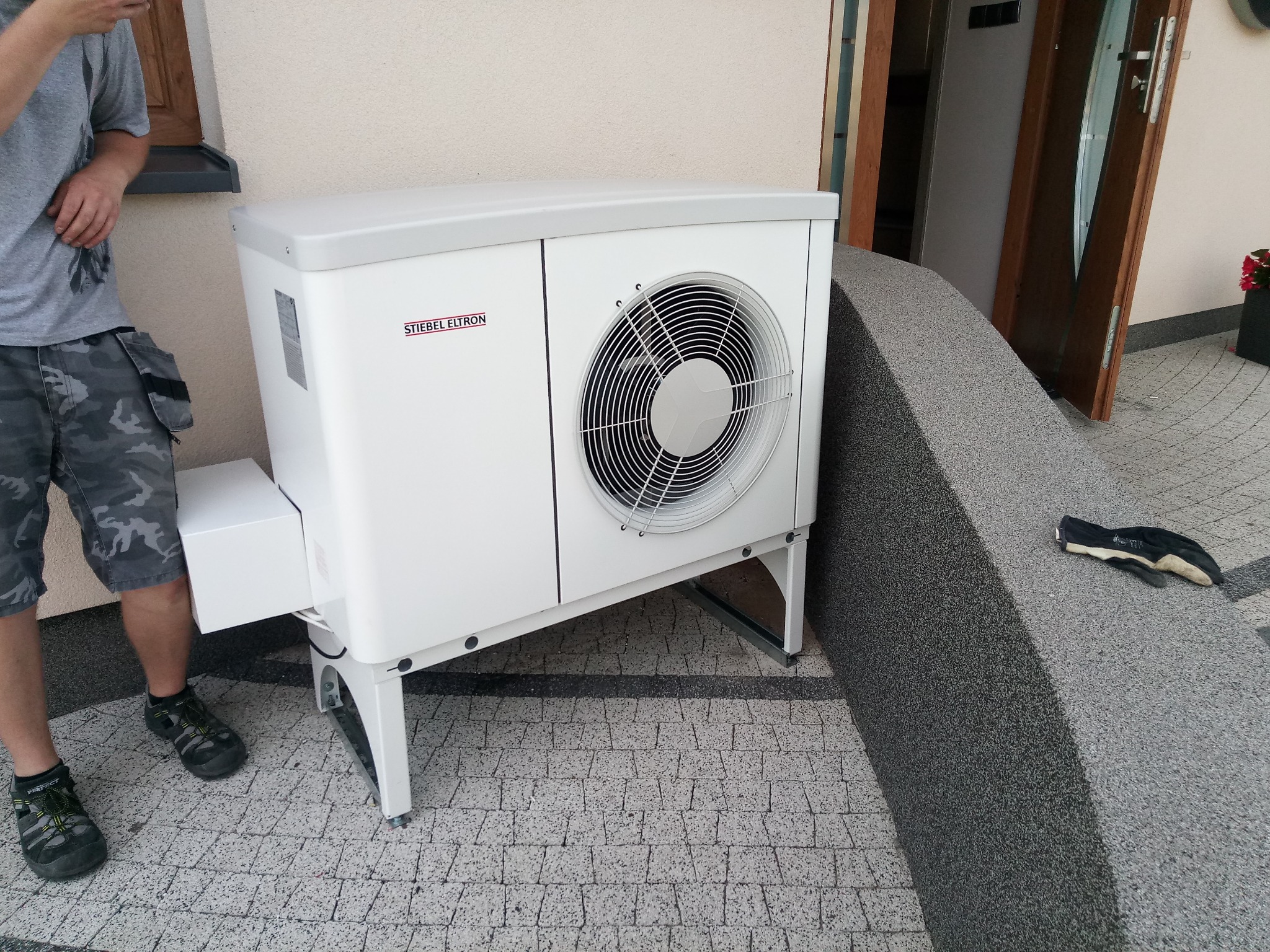 Biała pompa ciepła Stiebel Eltron zamontowana na kostce brukowej przed budynkiem, widoczne fragmenty osoby w szortach moro i robocze rękawice obok.