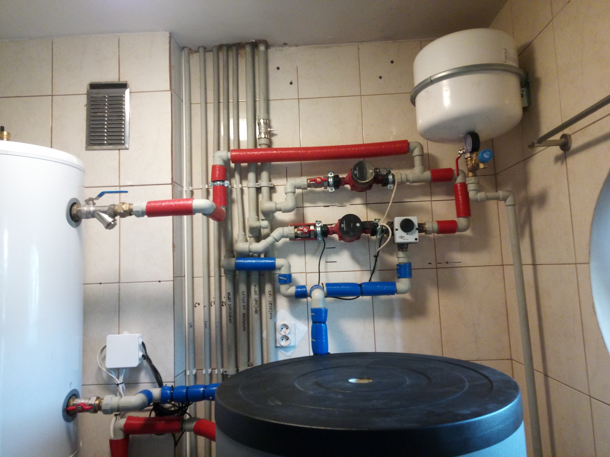 Kotłownia z widocznym zbiornikiem buforowym, zasobnikiem ciepłej wody użytkowej i rozbudowaną instalacją hydrauliczną z rurami w kolorach czerwonym i niebieskim, prowadzącymi do pompy obiegowej.