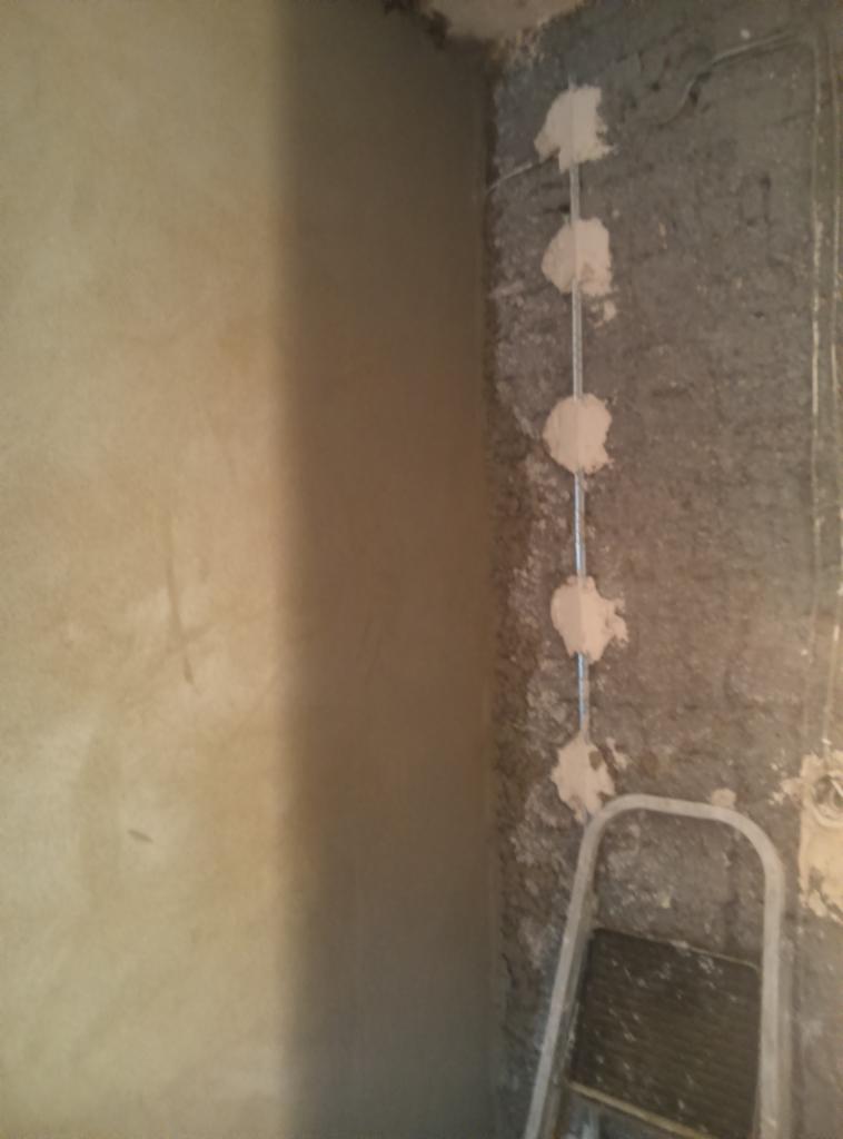 Fragment ściany: gładź szpachlowa po lewej, surowy mur z instalacją elektryczną po prawej, drabina w rogu. Etap prac wykończeniowych.