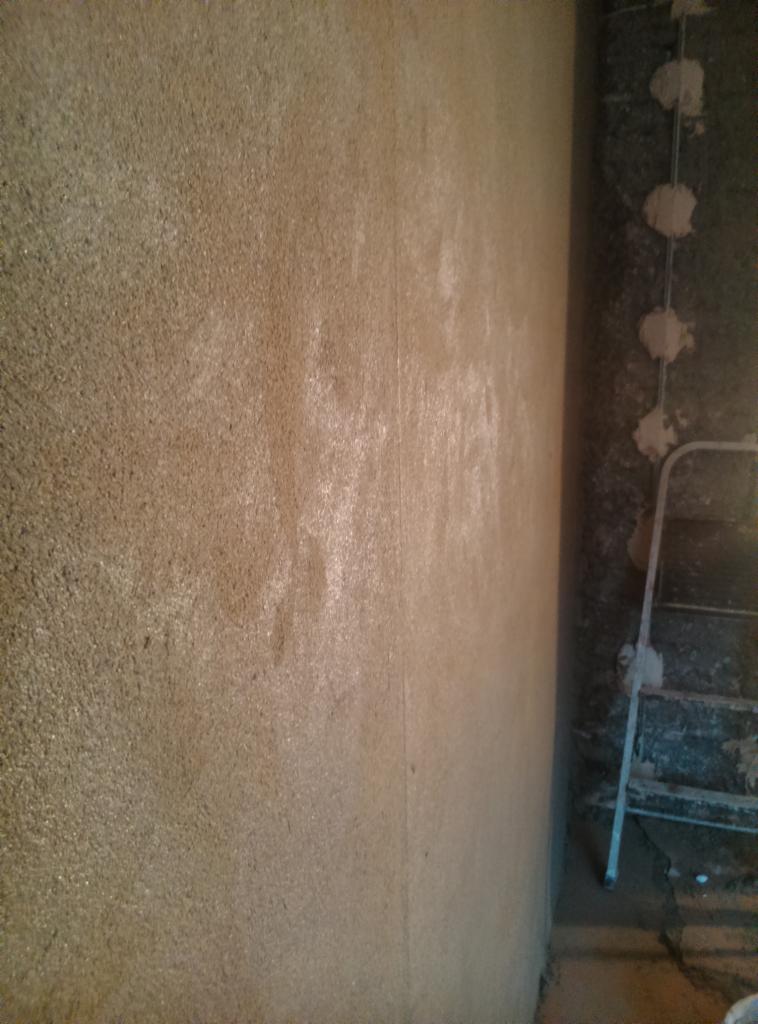 Ściana pokryta świeżym, beżowym tynkiem strukturalnym, widoczne delikatne cieniowanie i faktura, obok drabina malarska i fragment ściany z instalacją elektryczną w trakcie prac.