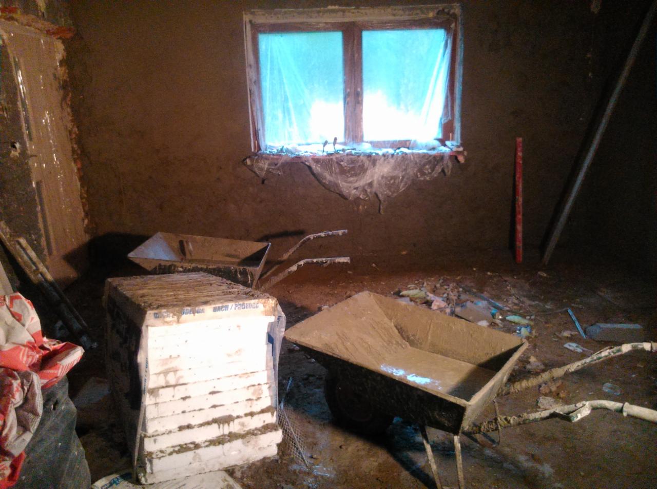 Wnętrze w trakcie tynkowania, widoczne taczki z zaprawą, stos płyt styropianowych, okno zasłonięte folią ochronną i świeżo otynkowane ściany.