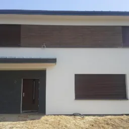 Dwukondygnacyjny dom z jasną elewacją, drewnianym akcentem na górze i szarym wykończeniem garażu. Okna z brązowymi roletami, teren wokół w stanie surowym.