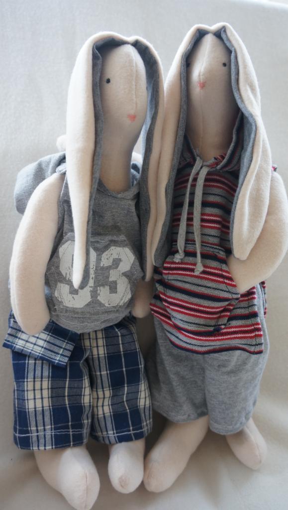 Dwa pluszowe króliki w ubraniach: jeden w szarej koszulce z numerem '93' i w niebieskich spodniach w kratę, drugi w bluzie w szaro-czerwone paski z kapturem i szarych spodniach.
