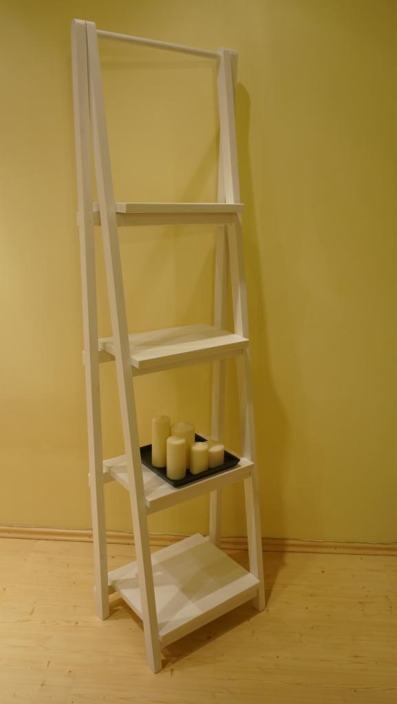 Biały, drewniany regał drabinkowy z trzema półkami, na jednej z nich czarny talerz z białymi świecami, ustawiony na drewnianej podłodze i tle żółtej ściany.