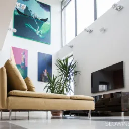 Jasny salon z wysokim sufitem, beżową sofą na metalowych nóżkach, telewizorem na ścianie, roślinami doniczkowymi i obrazami w nowoczesnym stylu.