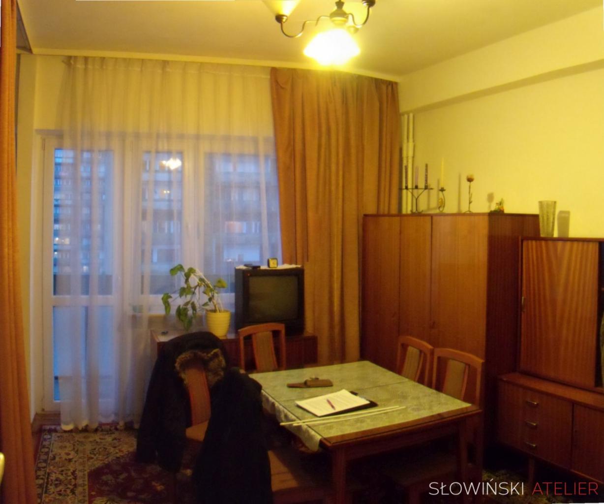 Pokój w Łodzi z meblami w stylu retro: stół nakryty zielonym obrusem, krzesła z drewnianymi oparciami, szafa i komoda z lat 70., telewizor kineskopowy na tle okna z firankami i zasłonami.