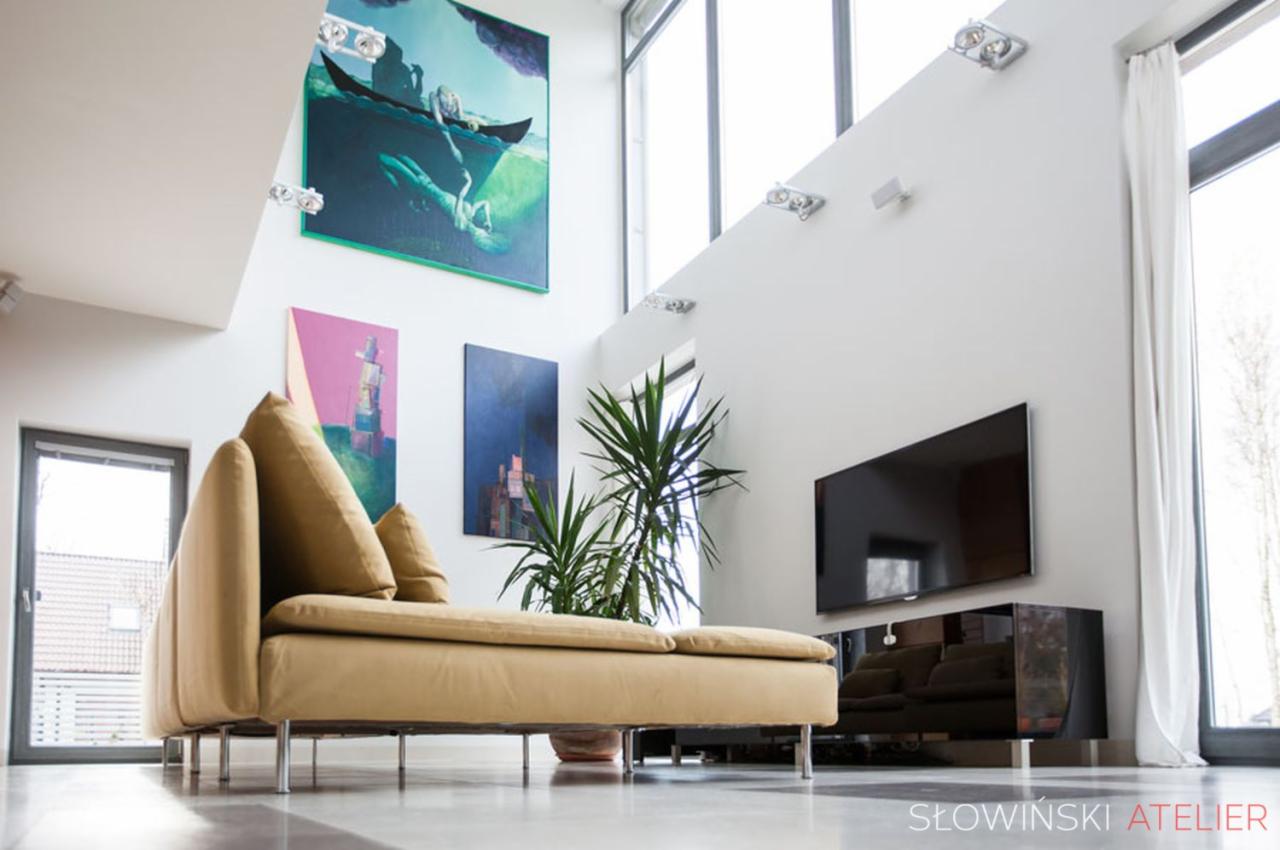 Jasny salon z wysokim sufitem, beżową sofą na metalowych nóżkach, telewizorem na ścianie, roślinami doniczkowymi i obrazami w nowoczesnym stylu.
