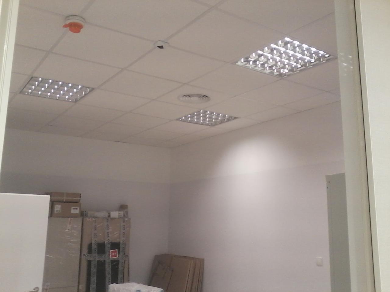 Wnętrze biurowe z zamontowanymi kwadratowymi lampami sufitowymi, czujnikiem dymu i kamerą monitoringu, widok przez futrynę drzwiową, w tle kartony ze sprzętem.