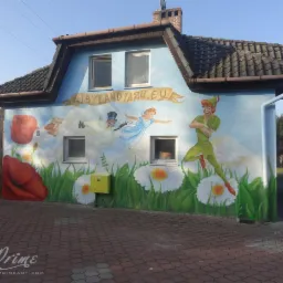 Budynek z muralem: Piotruś Pan, Dzwoneczek i kwiaty. Żółta skrzynka gazowa na ścianie. Widoczny dach i kostka brukowa. Nazwa na budynku: BYLANDIA24.EU.